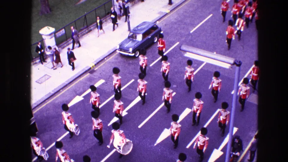 Elevated 1969 home-movie view of guardsmen in red tunics and dark bearskin-style hats marching past spectators on a city street.