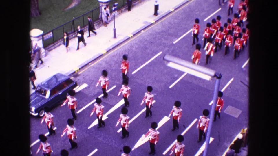 High-angle 1969 home-movie frame showing guardsmen spaced across the roadway, with a military band formation farther up the street.
