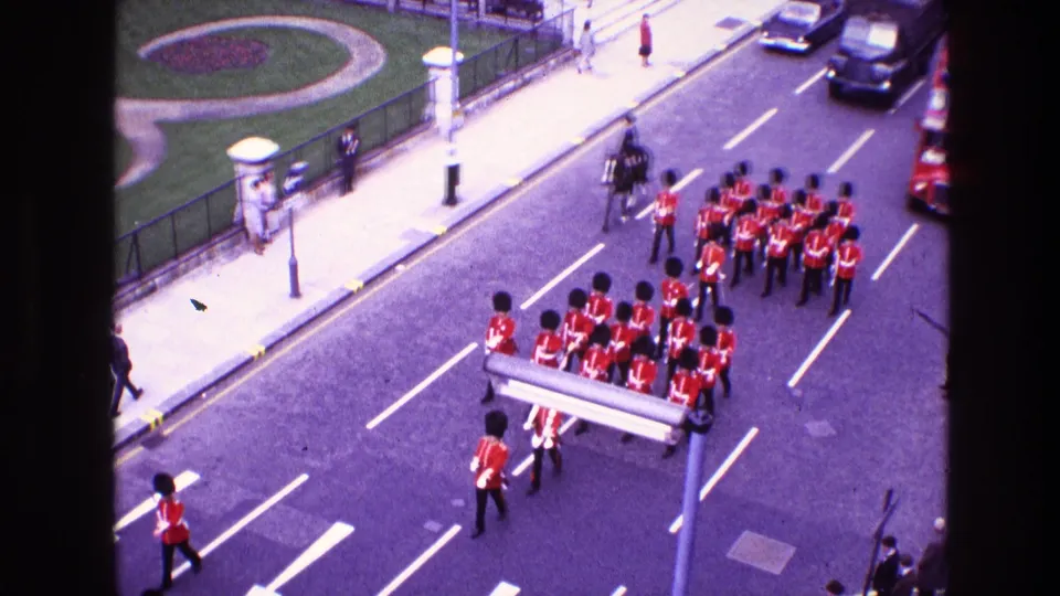 High-angle 1969 home-movie frame of a red-uniformed military band passing traffic, with a red double-decker bus partly visible.