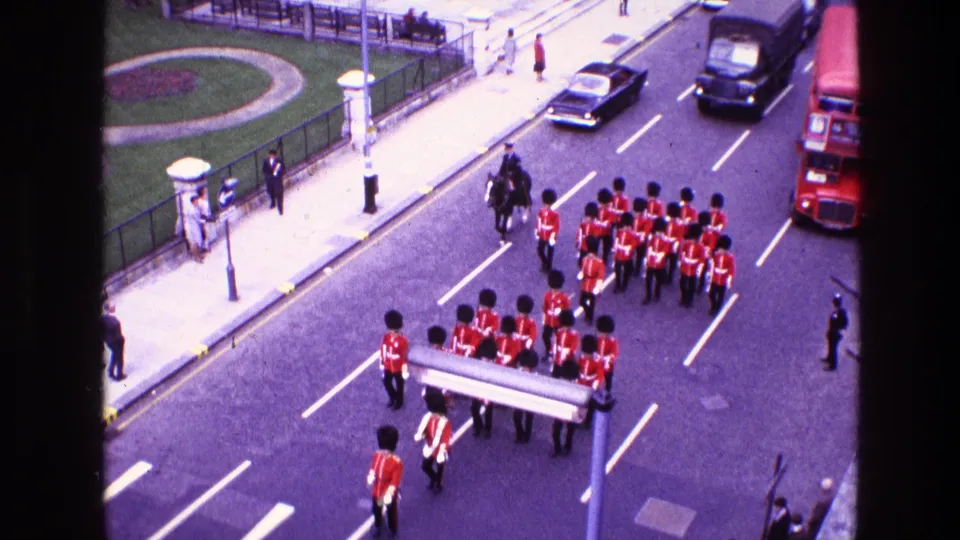1969 8mm frame of a ceremonial guard formation in red tunics and tall black hats marching on a city street with traffic nearby.