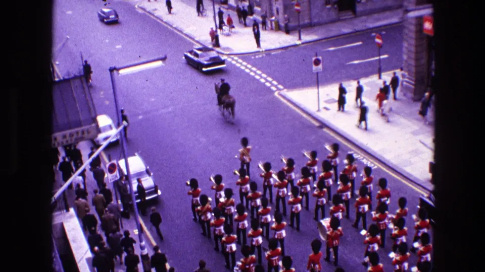 Elevated 8mm view of guards in red ceremonial uniforms moving through a city intersection with spectators, cars, and street signage visible.