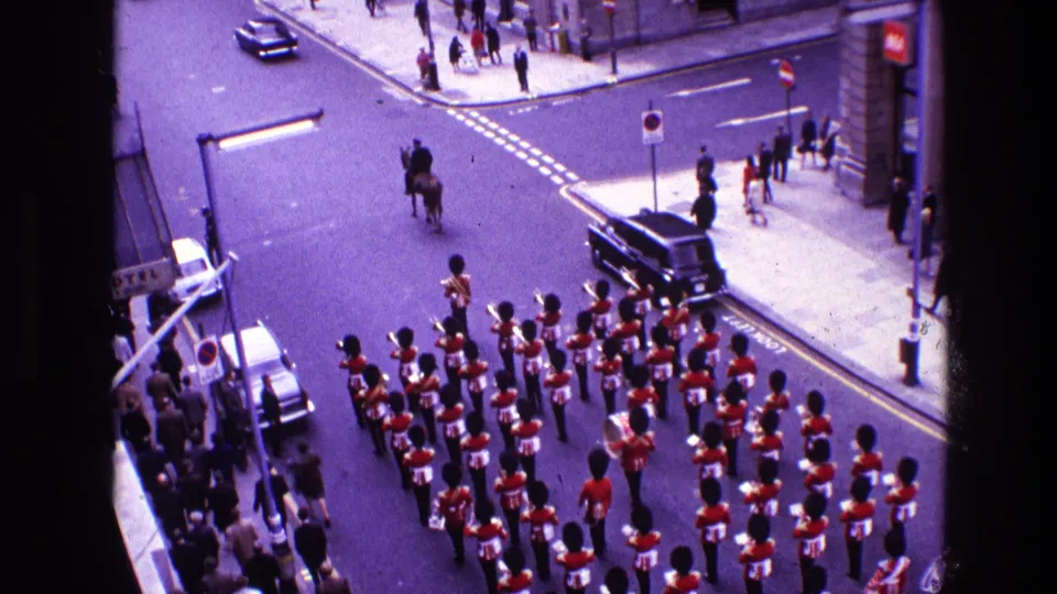 1969 home-movie frame showing red-uniformed guards with tall black hats marching past a dark car and sidewalk spectators on a city street.