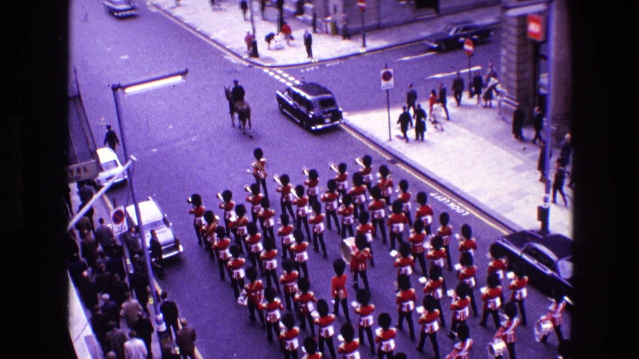 Marching band in red uniforms turns through a city street as pedestrians and cars pass nearby.