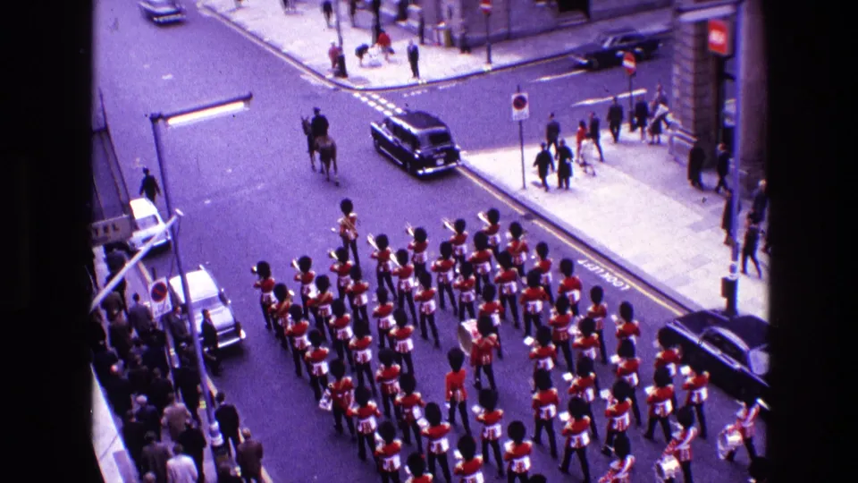 High-angle 1969 Ireland 8mm frame of a red-uniformed marching band moving through a city street with pedestrians, cars, and a mounted figure nearby.
