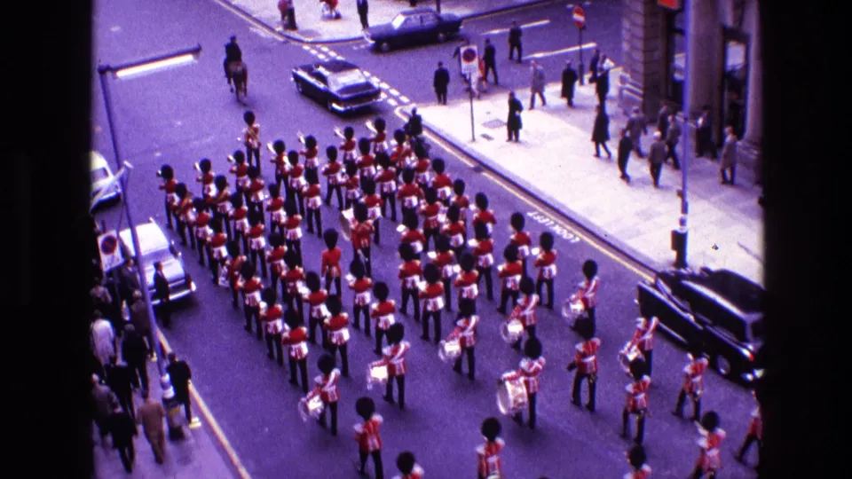 1969 Ireland 8mm frame showing the marching band from above, with red uniforms, drums or brass, a dark hearse-like vehicle, cars, and pedestrians.