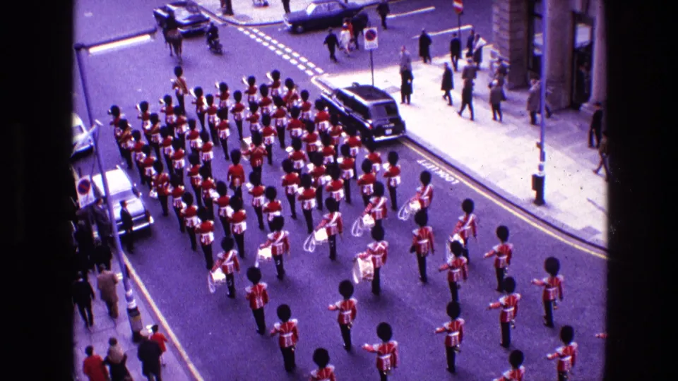High-angle 1969 Ireland home-movie frame of a marching band in red coats and black hats passing through a city street with cars and spectators.