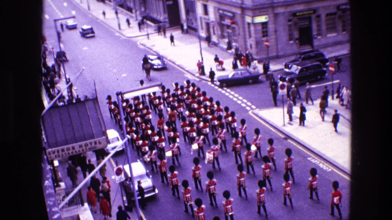 Marching band in red uniforms proceeds down a city street past spectators and parked cars.