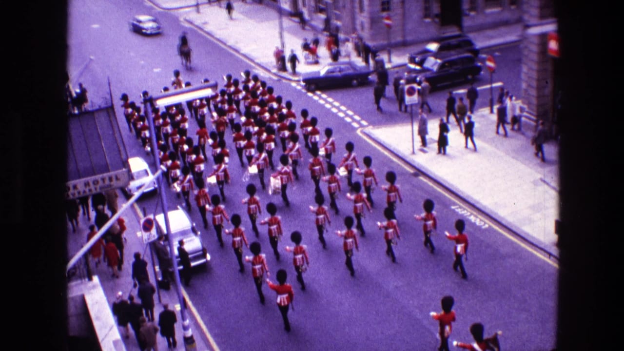 High-angle 1969 Ireland street parade view showing rows of red-uniformed band members, spectators, cars, and masonry buildings.