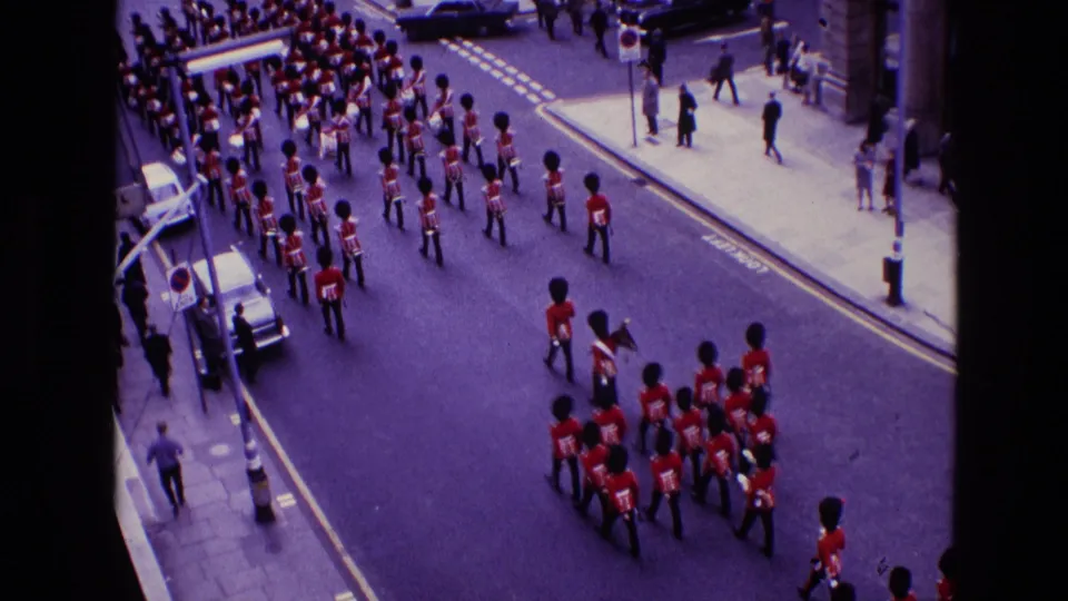 1969 Ireland home-movie frame with a red-uniformed marching band moving down a city street, seen from above with spectators nearby.