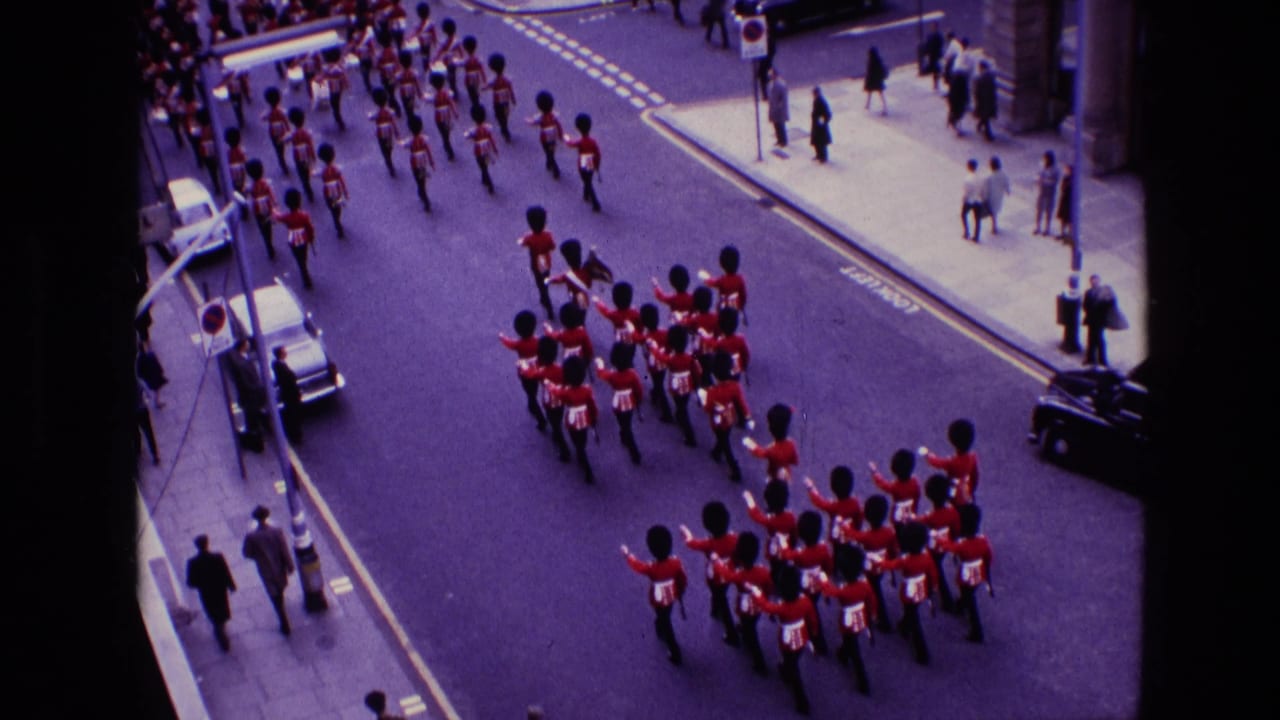 A formation of guards turns or advances along the street while pedestrians stand at the curb.