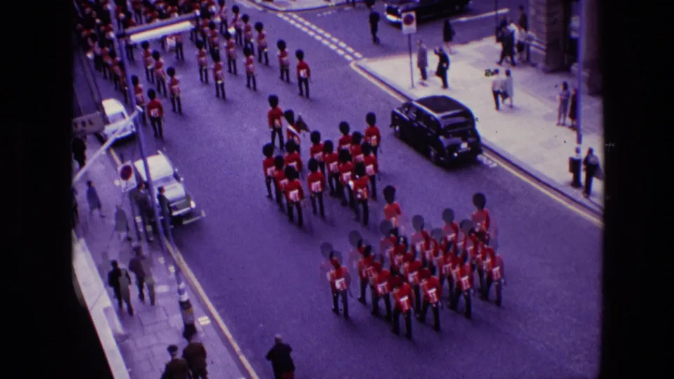 Elevated home-movie frame showing ceremonial guards in red uniforms marching through a London street scene in 1969.