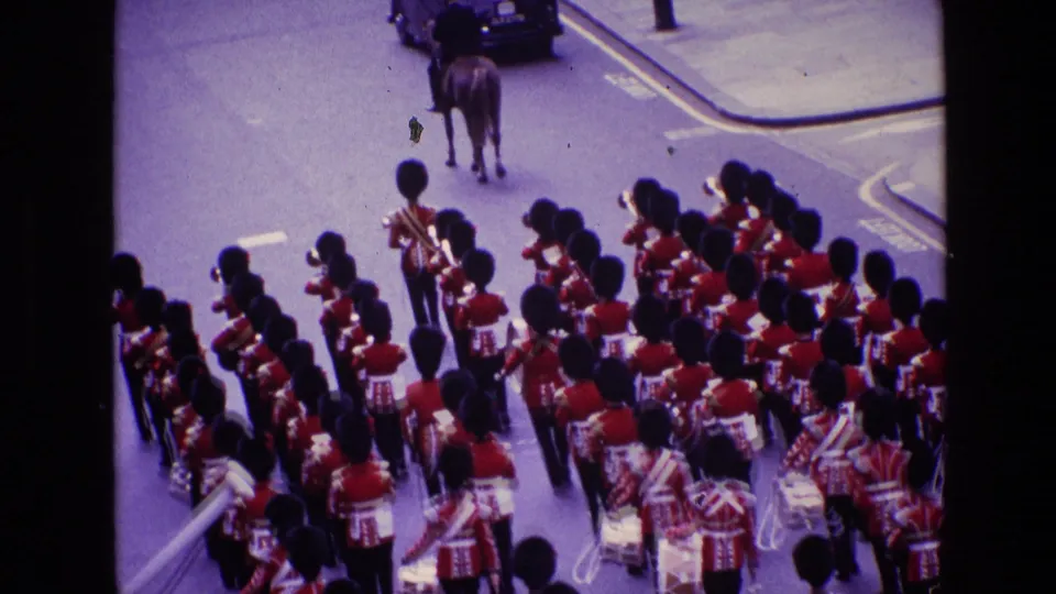 1969 Ireland home-movie frame showing drummers and guards in red ceremonial uniforms moving behind a mounted officer on a city street.