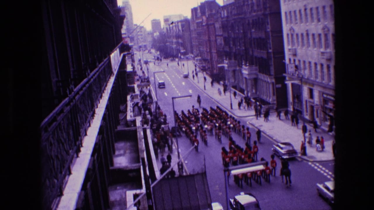 High-angle street view of a marching band in red uniforms passing between city buildings.
