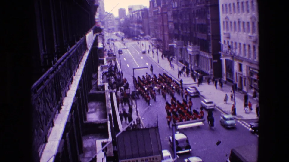1969 Ireland parade frame showing the red-uniformed group moving past an intersection, with spectators lining the pavement and cars parked or passing.