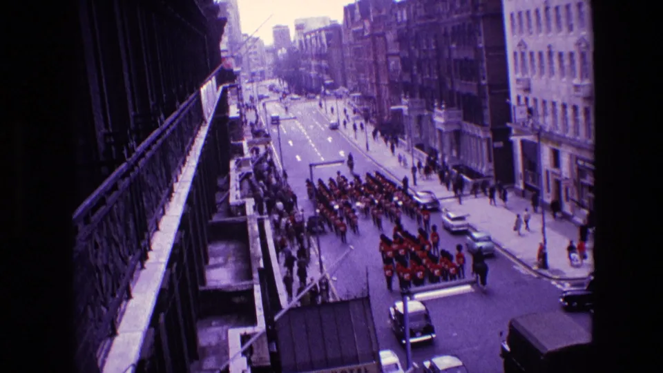 End frame from the 1969 Ireland 8mm clip, showing the civic parade receding along the street as cars and pedestrians occupy the foreground.