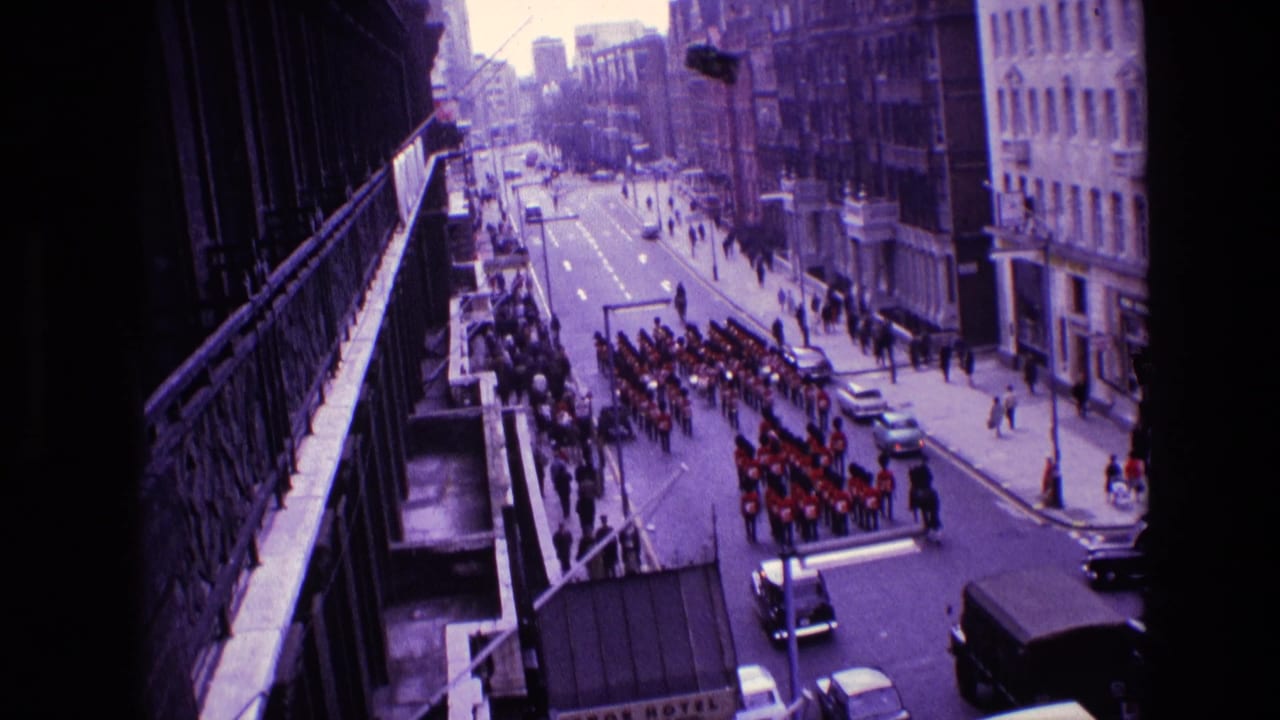 1969 Ireland 8mm frame showing a red-uniformed marching band parading along a city street beneath a balcony viewpoint.
