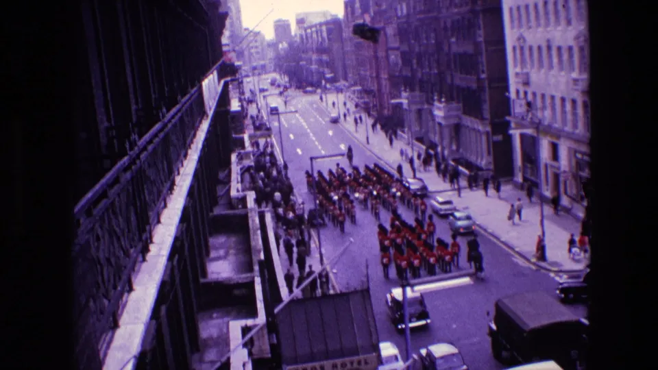 1969 Ireland 8mm frame showing a red-uniformed marching band parading along a city street beneath a balcony viewpoint.