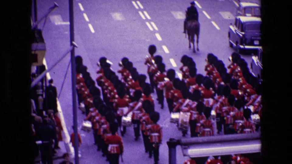1969 Ireland street parade frame showing a red-uniformed marching band following a rider on horseback, with parked cars at the curb.
