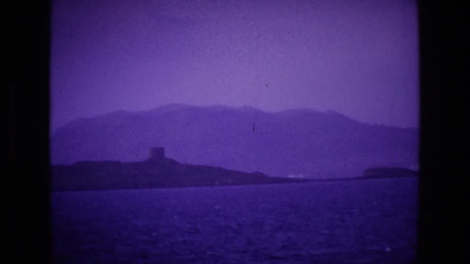 Wide 8mm frame of an Irish coastal view, with purple color shift, distant hills, a small tower, and sea in the foreground.