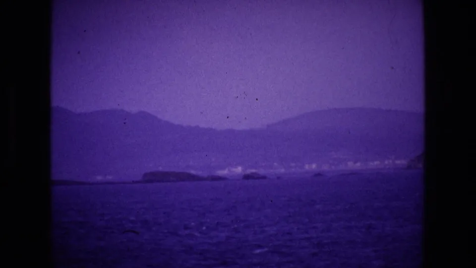 1969 Ireland 8mm frame showing a broad coastal view with open water, low shoreline, faint buildings, and hills beyond.