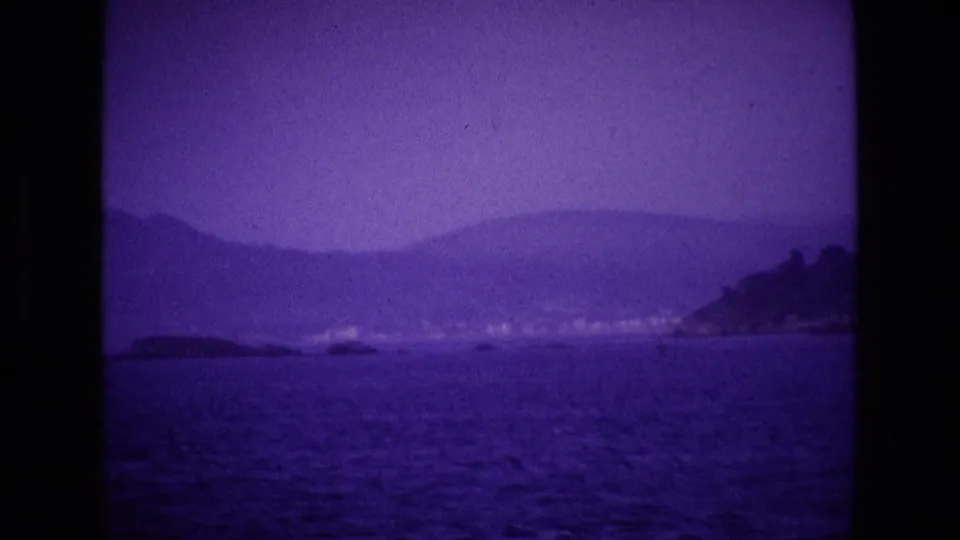 1969 Ireland 8mm frame of a coastal city or town seen from the water, with hills behind and a dark headland on the right.