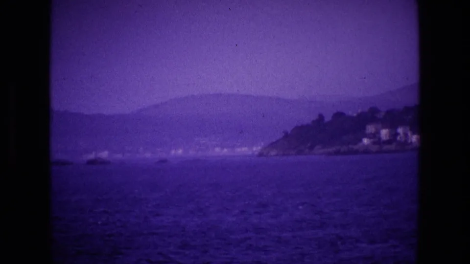 1969 8mm home-movie frame from Ireland showing blue water, hazy hills, and distant white buildings along the coast.