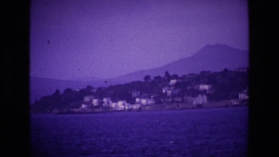 1969 Ireland 8mm frame of white coastal buildings below mist-covered mountains, seen from a moving boat.