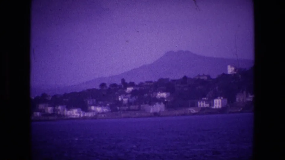 1969 8mm home-movie frame of an Irish coastal shoreline across water, with pale buildings below a mountain ridge.