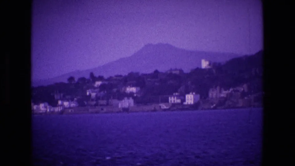 Purple-shifted 1969 Irish coastal view across water, showing shoreline buildings and a broad mountain form behind them.