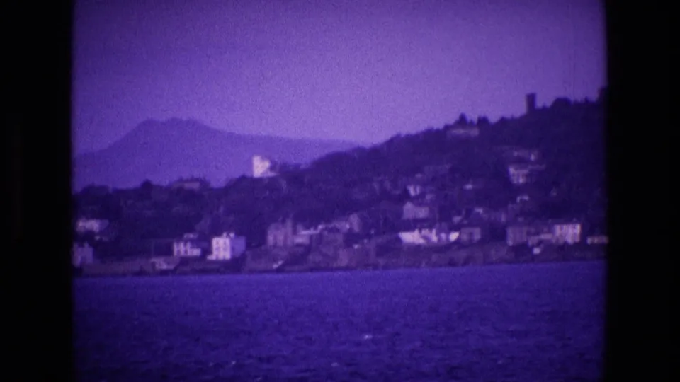 1969 8mm frame from Ireland showing water in the foreground, hillside buildings, and a softened mountain shape to the left.