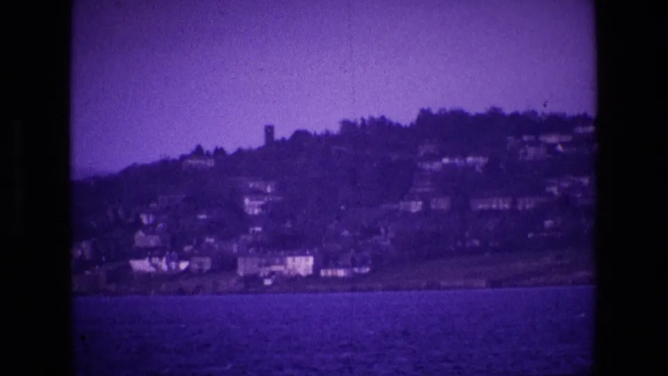 Late-1960s Irish home-movie view of a coastal hillside and waterfront buildings, tinted purple by the film image.