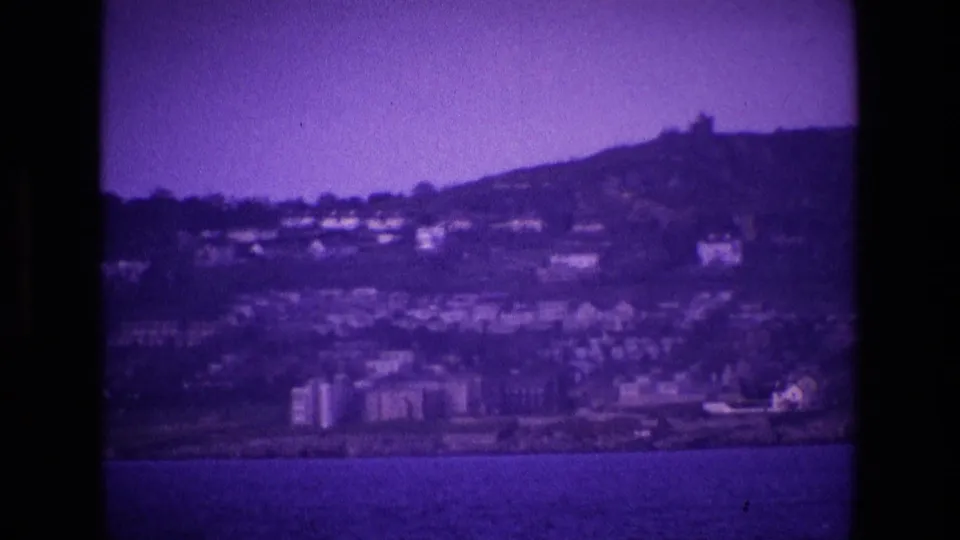 Purple-tinted 8mm frame showing an Irish seaside hillside with homes, low buildings, and calm water in the foreground.