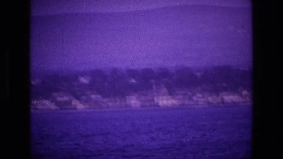 1969 Ireland home-movie frame showing open water and a distant shoreline with buildings and trees, viewed from a moving boat.
