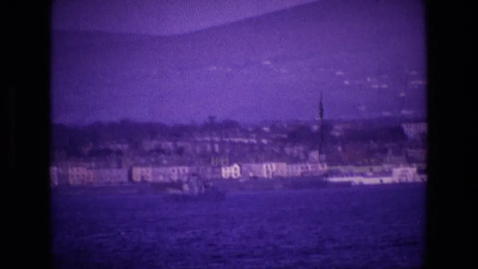 Ireland coastal home-movie frame showing water, distant buildings, trees, and a tall spire-like feature under a purple 8mm cast.