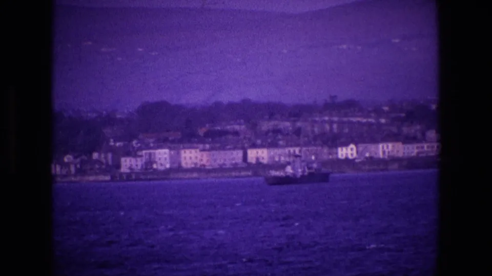 1969 Ireland 8mm frame with a small boat crossing open water in front of a distant shoreline and waterfront buildings.