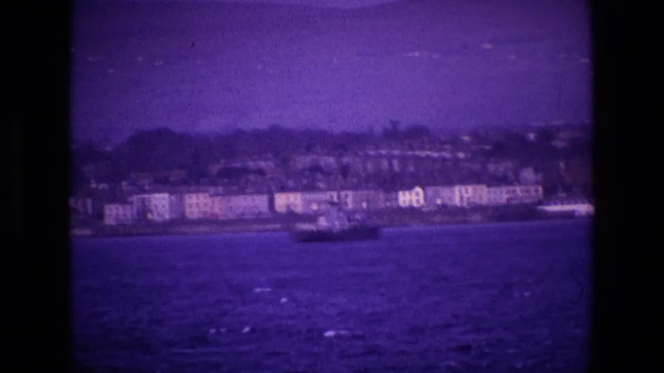1969 Ireland home-movie frame showing a shoreline town and small vessel viewed from water with a strong purple film cast.