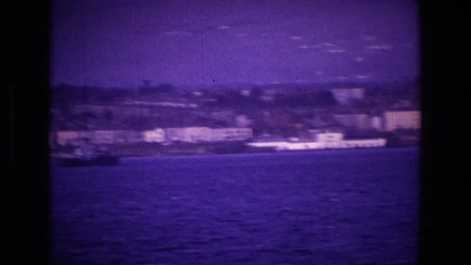 Purple-tinted 8mm view of an Irish waterfront with a long white vessel near the shore, filmed from the water in 1969.