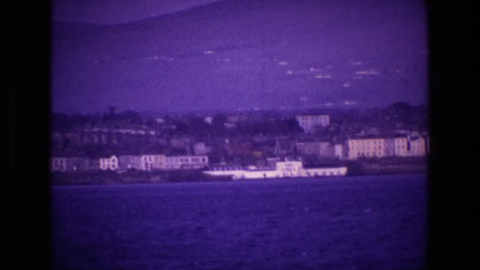 1969 Ireland 8mm frame showing a coastal town across the water, a long white vessel, and low hills under a purple sky.