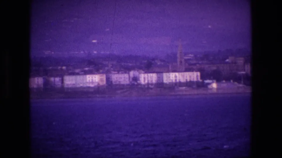1969 8mm Ireland coastal view with waterfront buildings and a distant church steeple across the water.