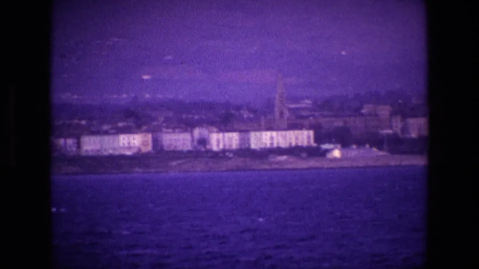 Final 1969 Ireland 8mm frame of a coastal town across the water, with a church steeple and hazy hills.