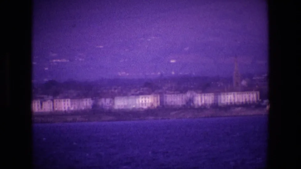 1969 Ireland home-movie frame showing a distant coastal town, pale waterfront buildings, and a church spire across dark water.