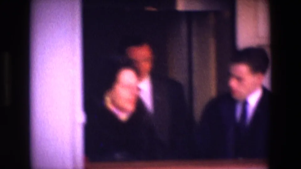 Blurry 1969 home-movie interior showing adults in dark dress or suits standing close together near curtains.