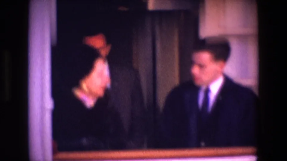 Dim, purple-tinted interior frame with two adults in dark suits or formal clothing standing near a curtained doorway.