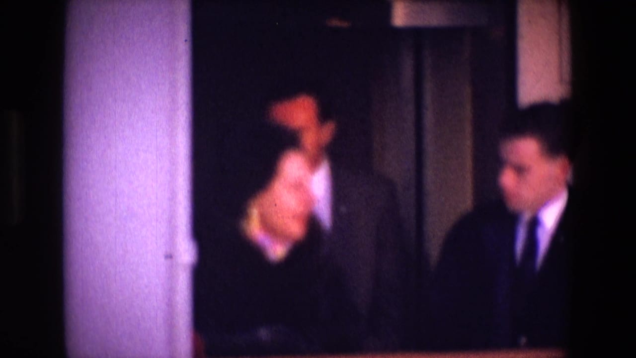 Two men in dark suits stand near a woman inside a dimly lit building.