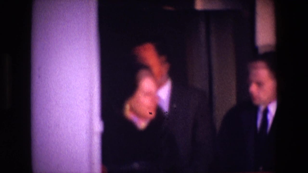 A woman and two suited men stand near a doorway in a dark interior.