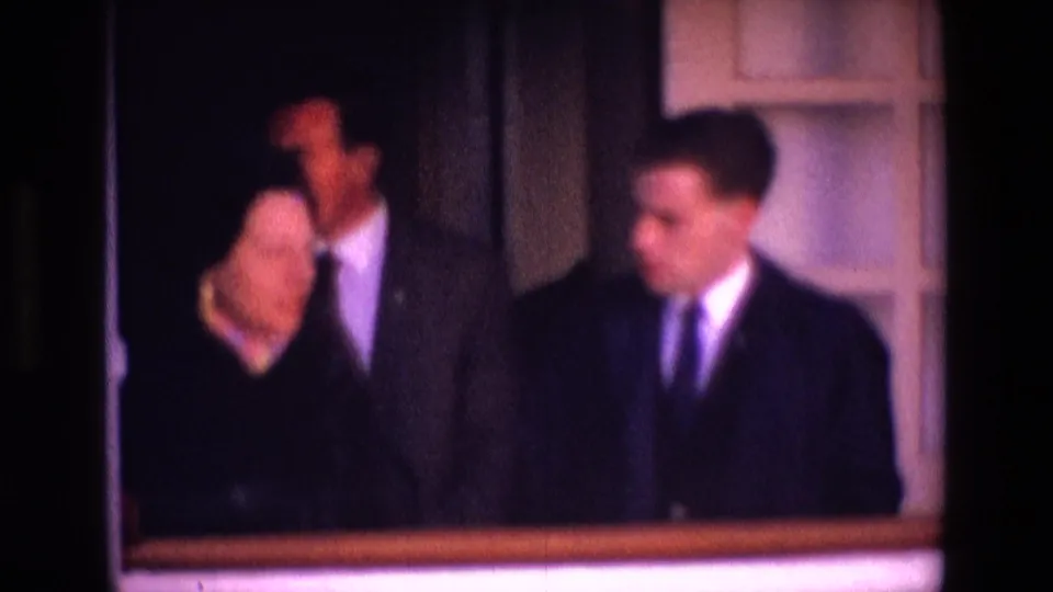 In the 1969 Ireland clip, the woman and suited men appear behind a low railing or sill within a softly lit interior.
