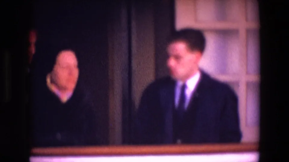 In 1969 Ireland home-movie footage, two adults stand in a dim interior, one wearing a suit and tie beside a woman in dark clothing.