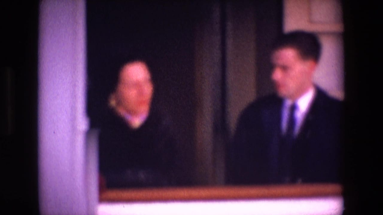The frame shows two adults indoors in Ireland, with a suited man turned toward a woman near white wall trim and a dark doorway.