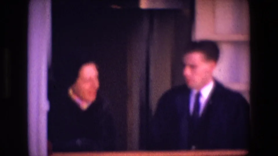 A soft 8mm frame shows two adults in a dim Irish interior, with the man in a dark suit and the woman beside white trim.