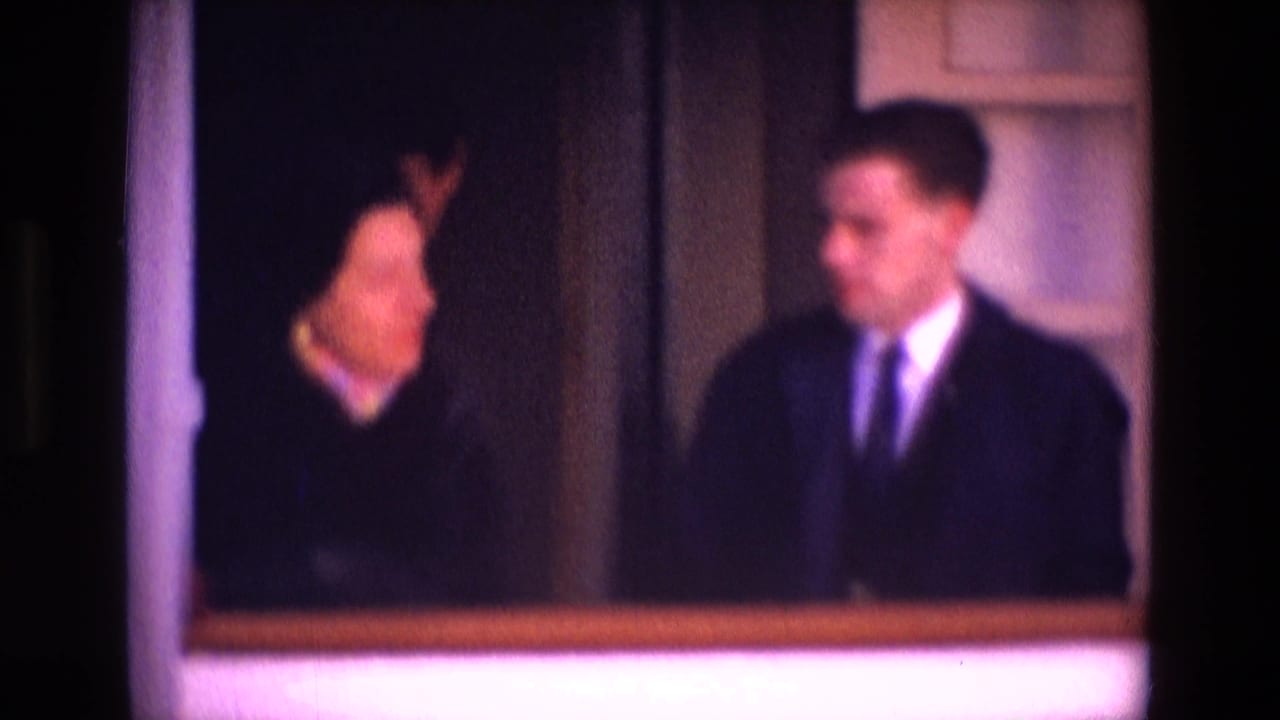 Woman in dark clothing and man in a suit standing near a boat window or doorway.